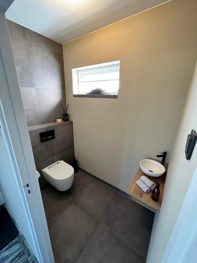 Gastentoilet met hangend toilet en kleine houten wastafel onder een raam